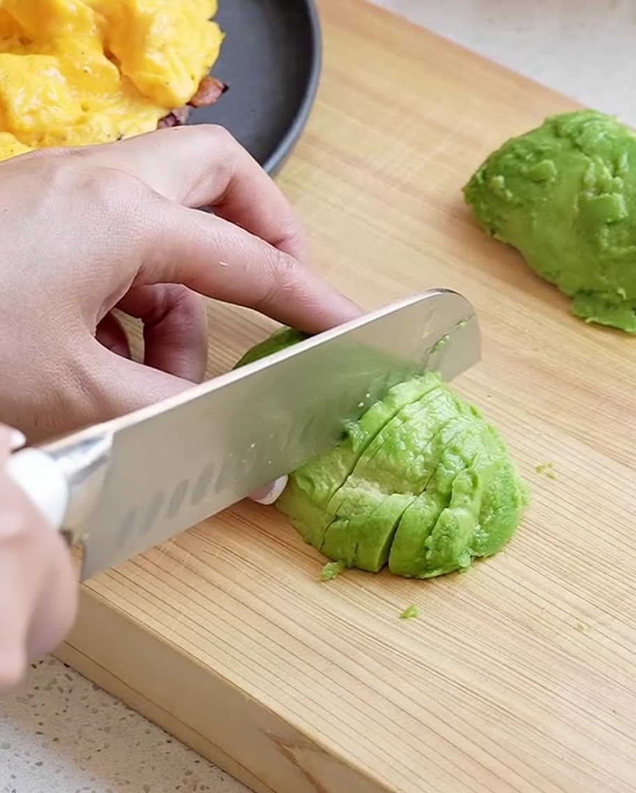 Una mano che usa un coltello da cucina per affettare una metà di avocado sbucciato in pezzi sottili e puliti su un tagliere di legno.