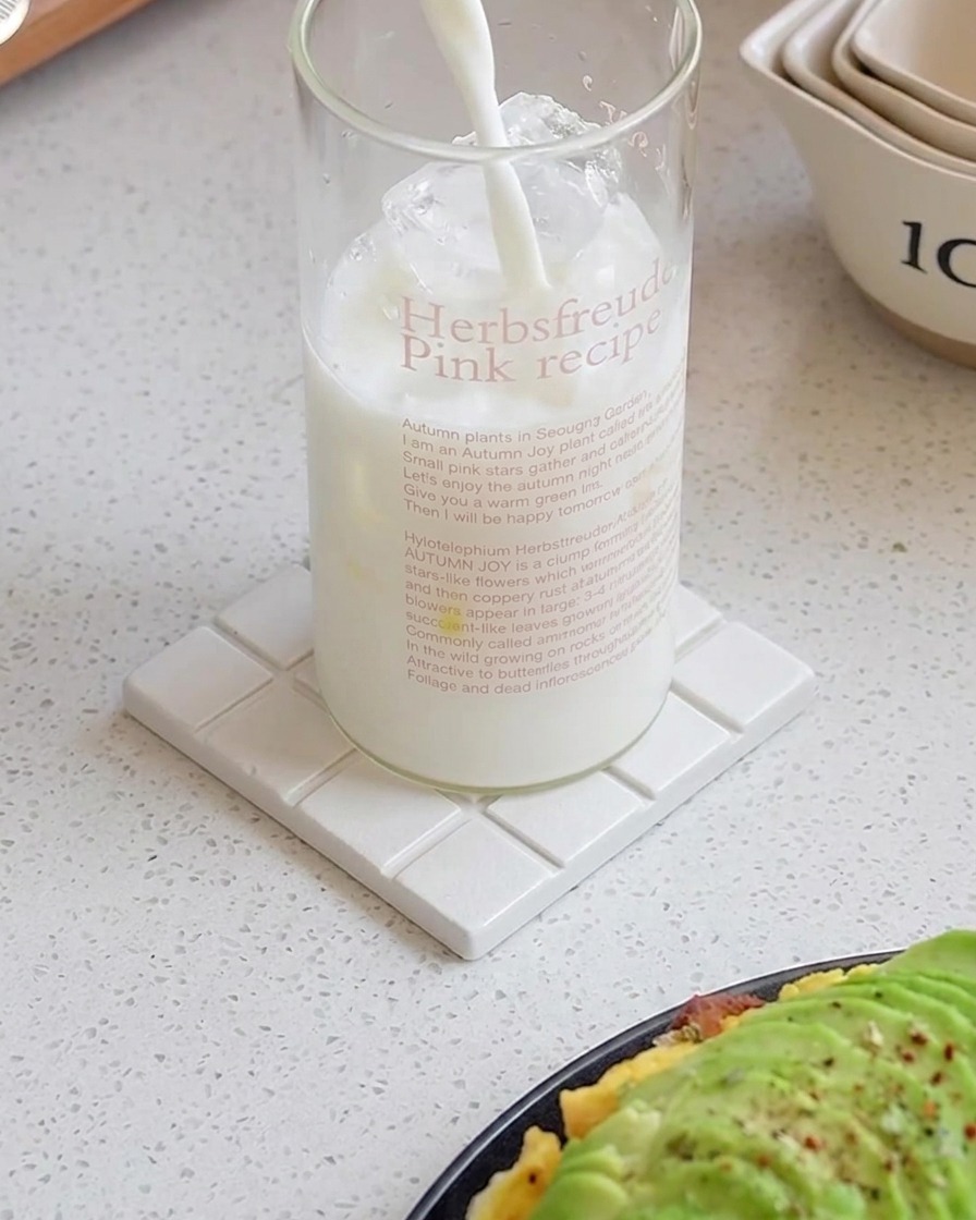 Latte versato in un bicchiere alto pieno di cubetti di ghiaccio su un sottobicchiere bianco.