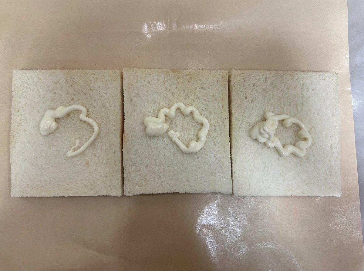 Tre fette di pane bianco senza crosta disposte una di fianco allaltra su carta da forno marrone, ciascuna guarnita con una linea ondulata di salsa bianca.