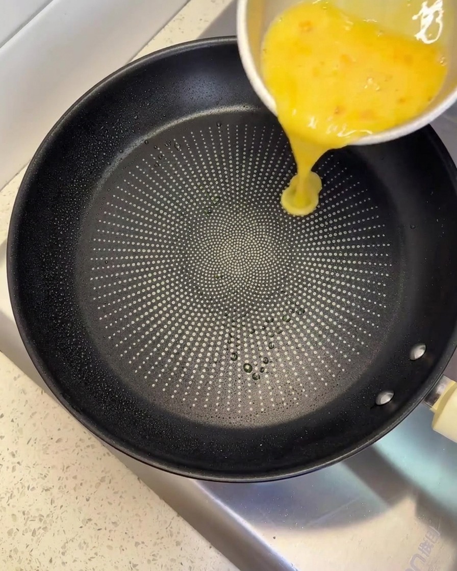 Versamento del composto di uova sbattute da una ciotola di metallo in una padella antiaderente nera.