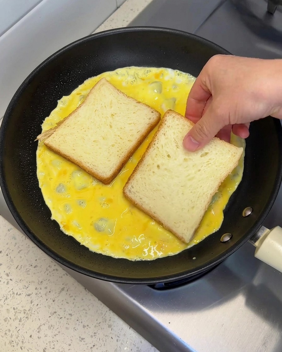 Una mano che posiziona due fette di pane bianco una accanto allaltra sopra luovo ancora liquido in padella.