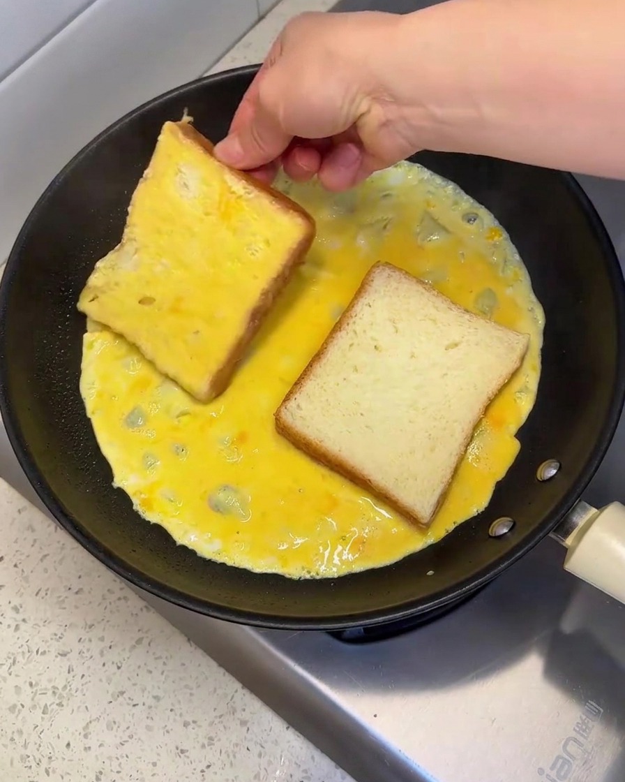 Una mano che posiziona e gira una fetta di pane in una padella calda piena di uova sbattute crude per coprire entrambi i lati.