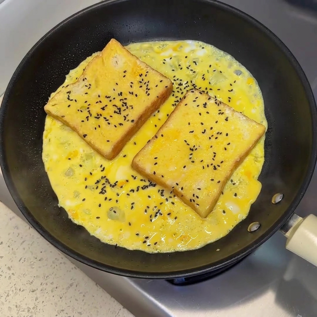 Semi di sesamo nero cosparsi su due fette di pane adagiate su una frittata a metà cottura in padella.