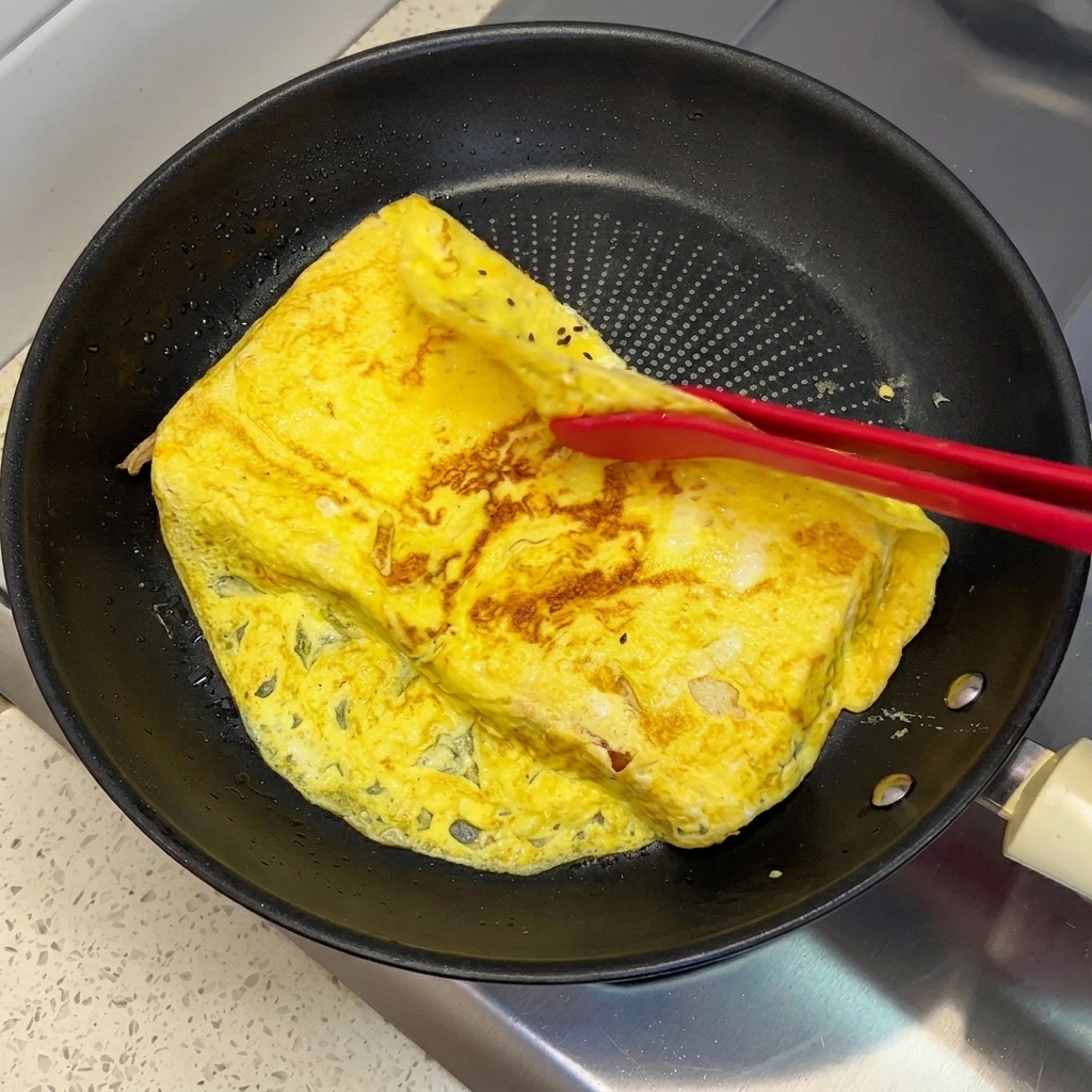 Pinze in silicone rosso che piegano i bordi sporgenti di una frittata cotta sopra due fette di pane in padella.