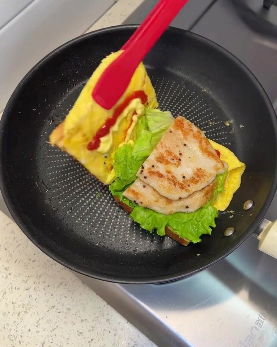 Un paio di pinze rosse che sollevano e piegano un lato del pane per chiudere il panino in padella.