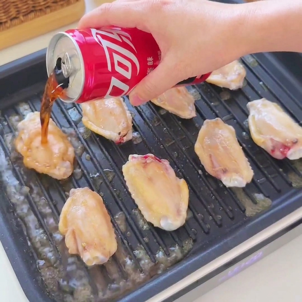 Cola e salsa di soia versate da una lattina rossa sulle ali di pollo dorate che sfrigolano in una bistecchiera nera.