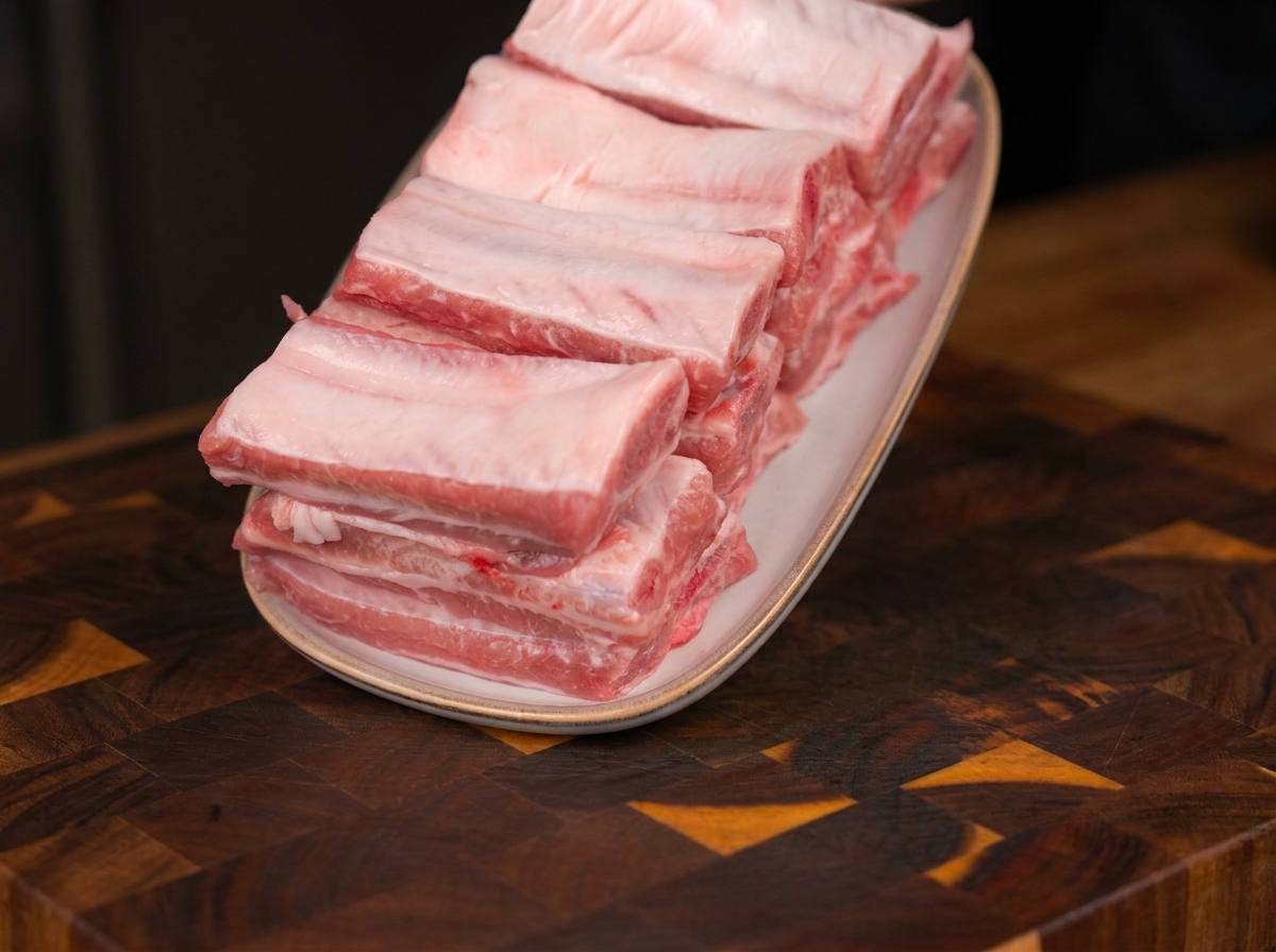Costine di maiale fresche e crude disposte ordinatamente su un piatto ovale bianco, appoggiato su un tagliere di legno.
