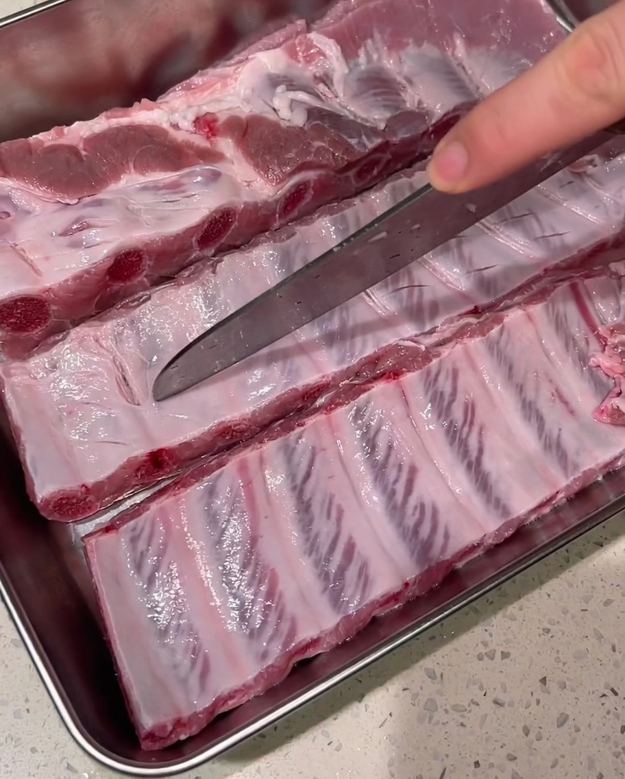 Un coltello affilato che pratica tagli superficiali sulla carne delle costine crude su un vassoio di metallo.