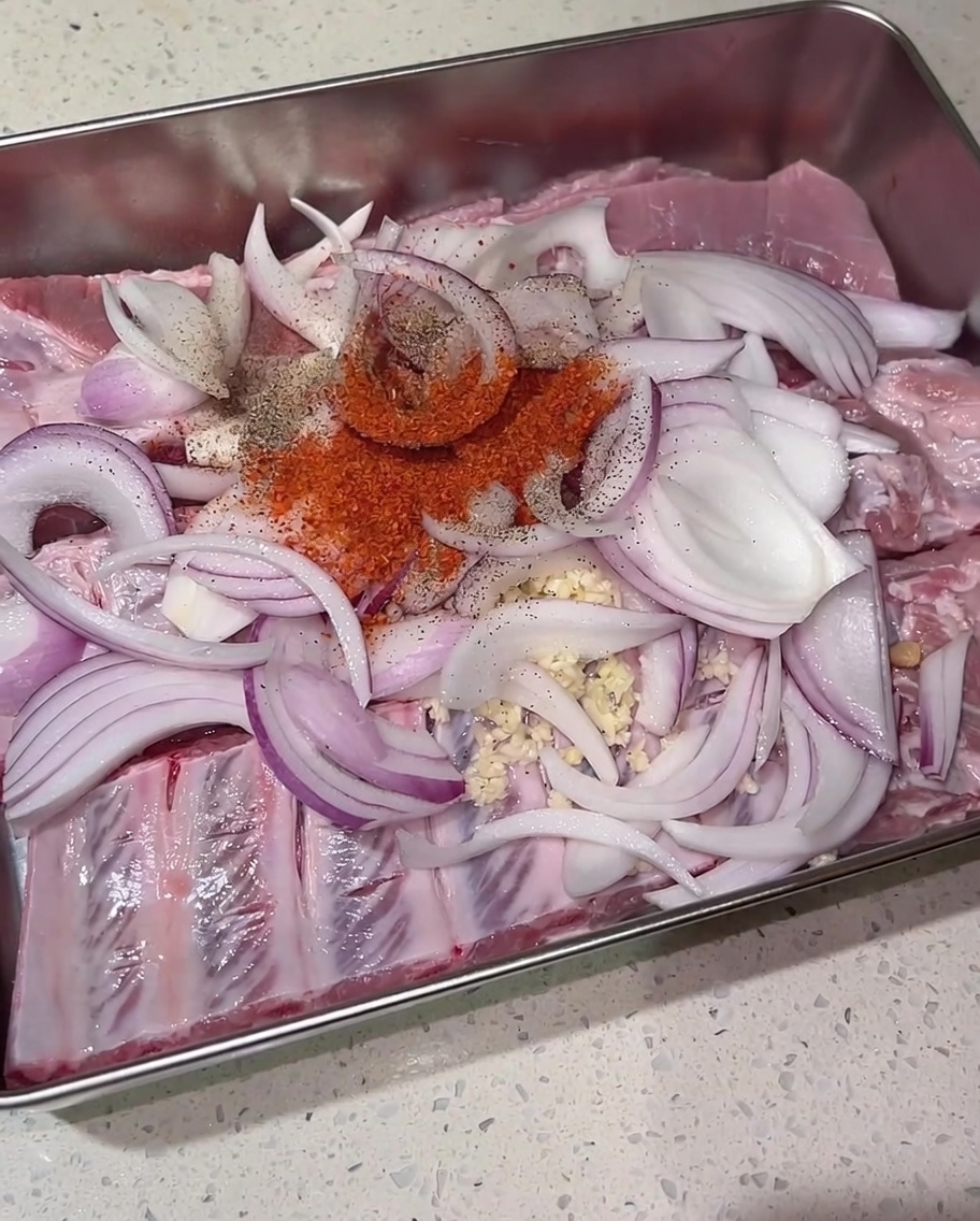 Un mucchietto di peperoncino in polvere, pepe nero e zucchero di canna adagiati sopra le costine crude e le cipolle in un vassoio di metallo.