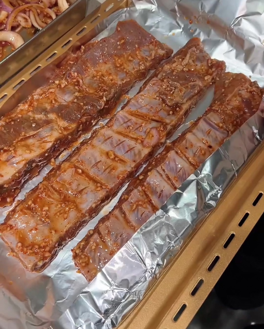 Tre file di costine marinate adagiate su un foglio di alluminio sopra una teglia dorata, pronte per essere avvolte.