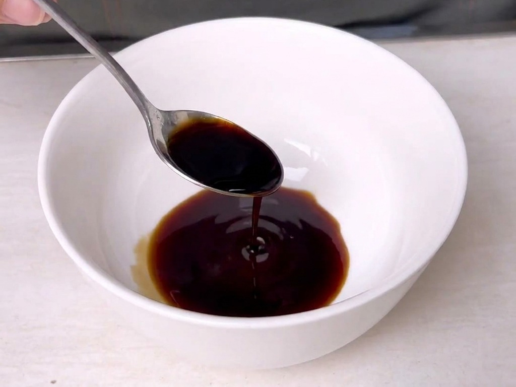 Un cucchiaio di metallo mescola una salsa scura in una ciotolina di ceramica bianca.