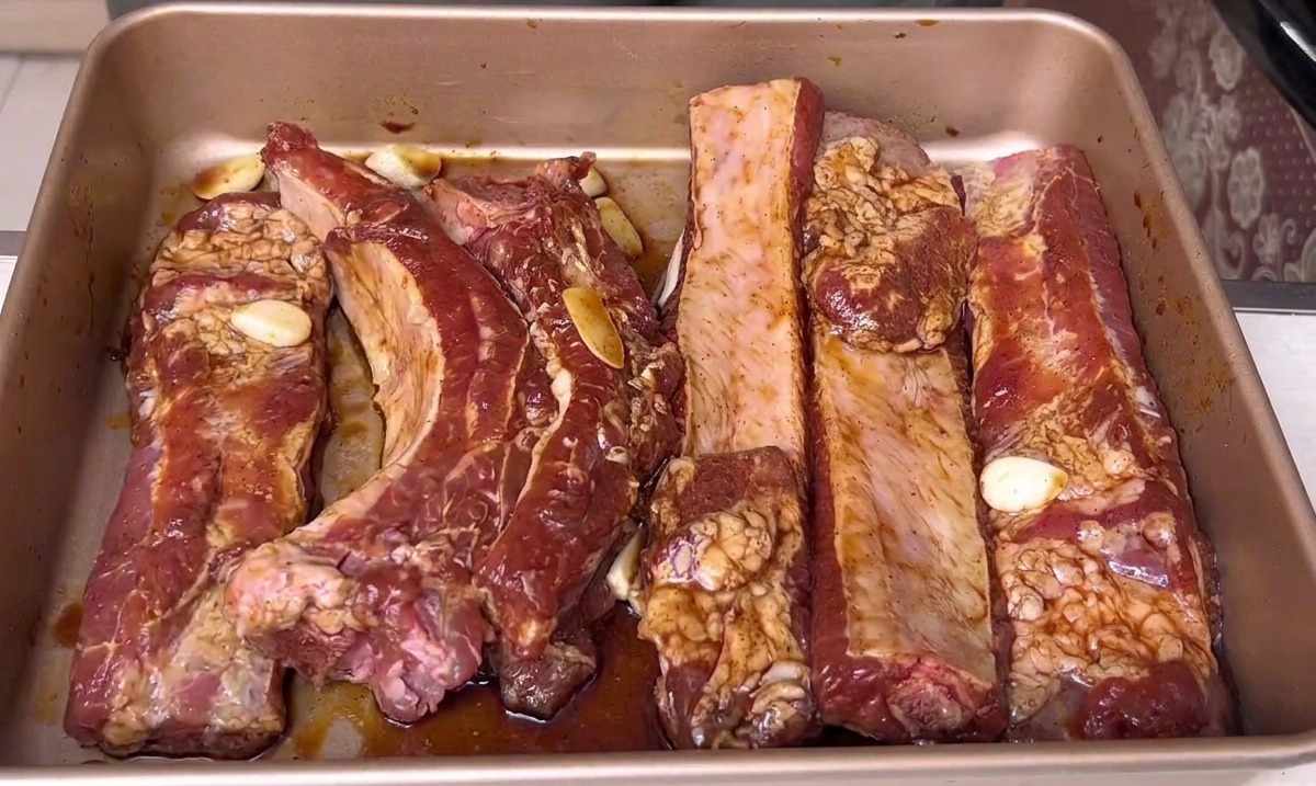 Costine di maiale marinate che riposano in una teglia dorata, pronte per essere infornate.