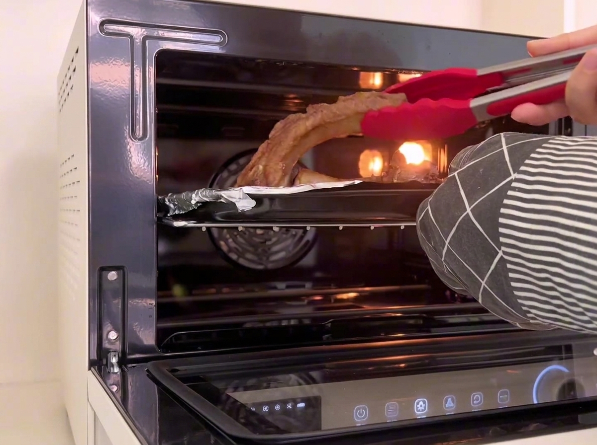 Pinze in silicone rosso girano dei pezzi di costine di maiale allinterno della teglia nel forno.