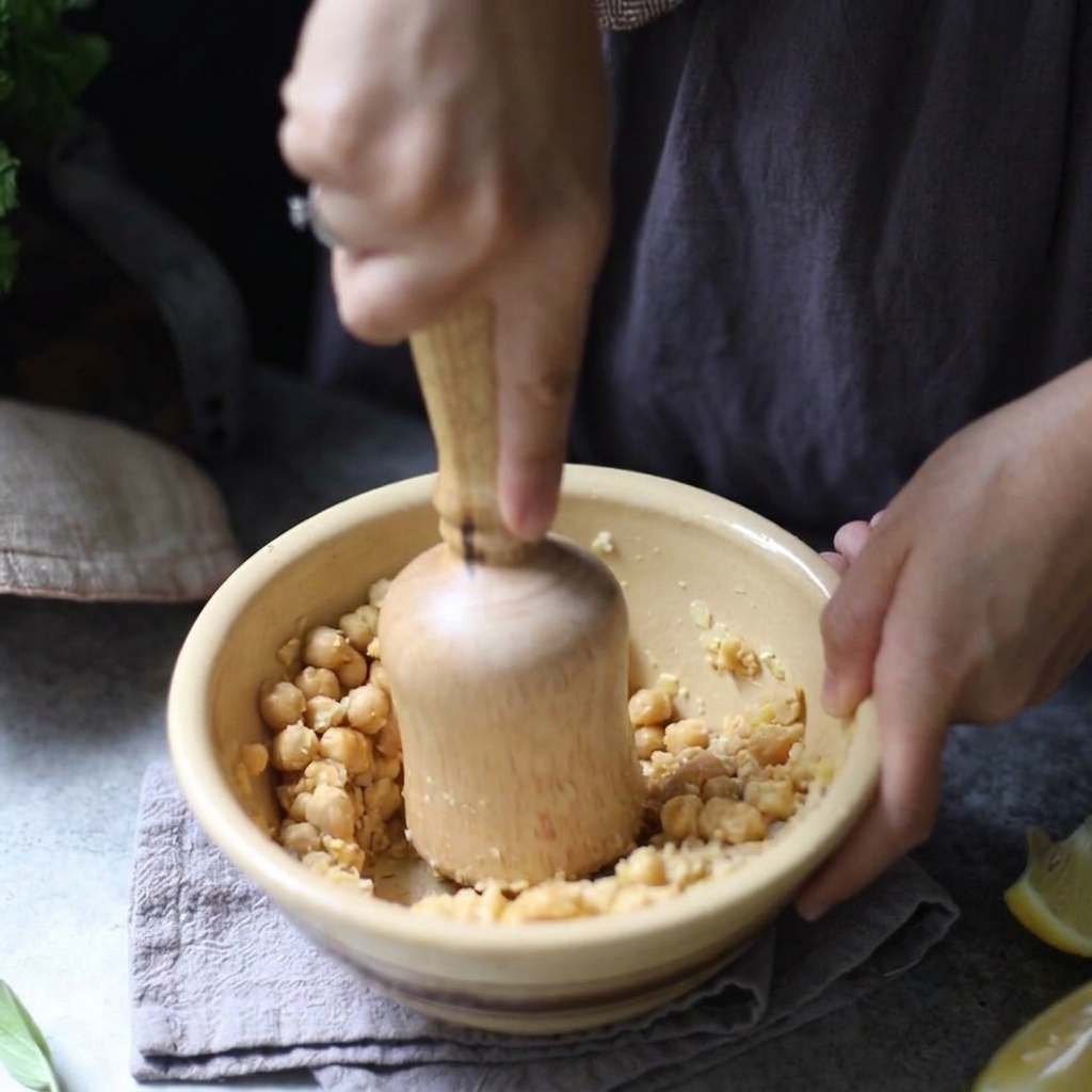 Una persona usa un pestello di legno per schiacciare manualmente i ceci bolliti in una ciotola di ceramica gialla.