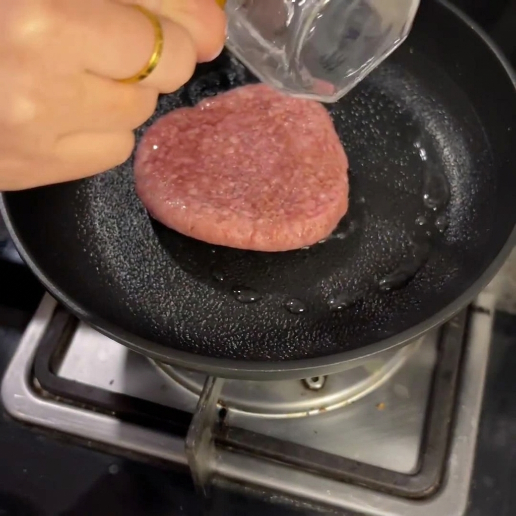 Un hamburger di manzo crudo in cottura dentro una padella con una piccola quantità di liquido, coperto da un coperchio.