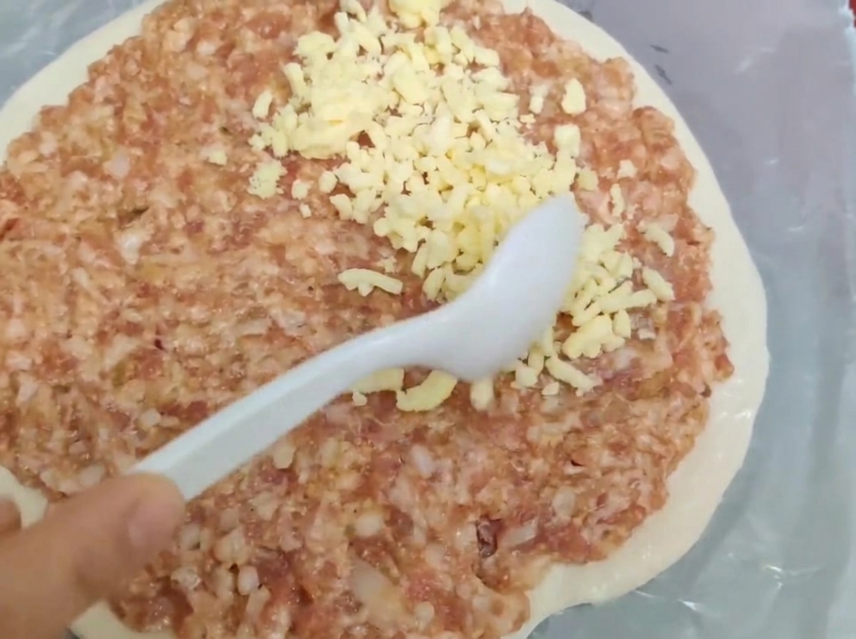 Un cucchiaio bianco che spalma carne macinata cruda e formaggio grattugiato su una base di pasta rotonda.