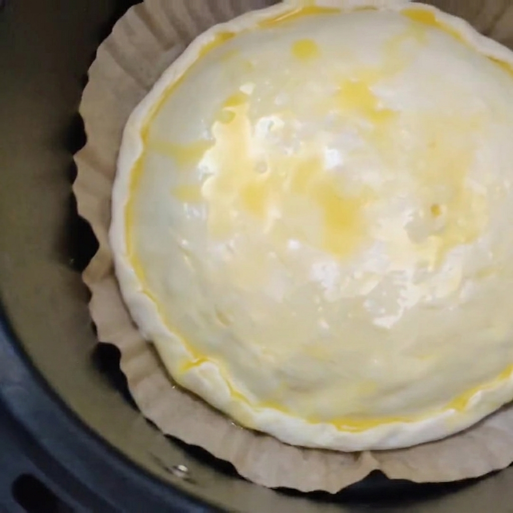 Una torta di carne e formaggio non cotta con la superficie spennellata duovo che riposa su carta da forno marrone allinterno del cestello nero della friggitrice ad aria.