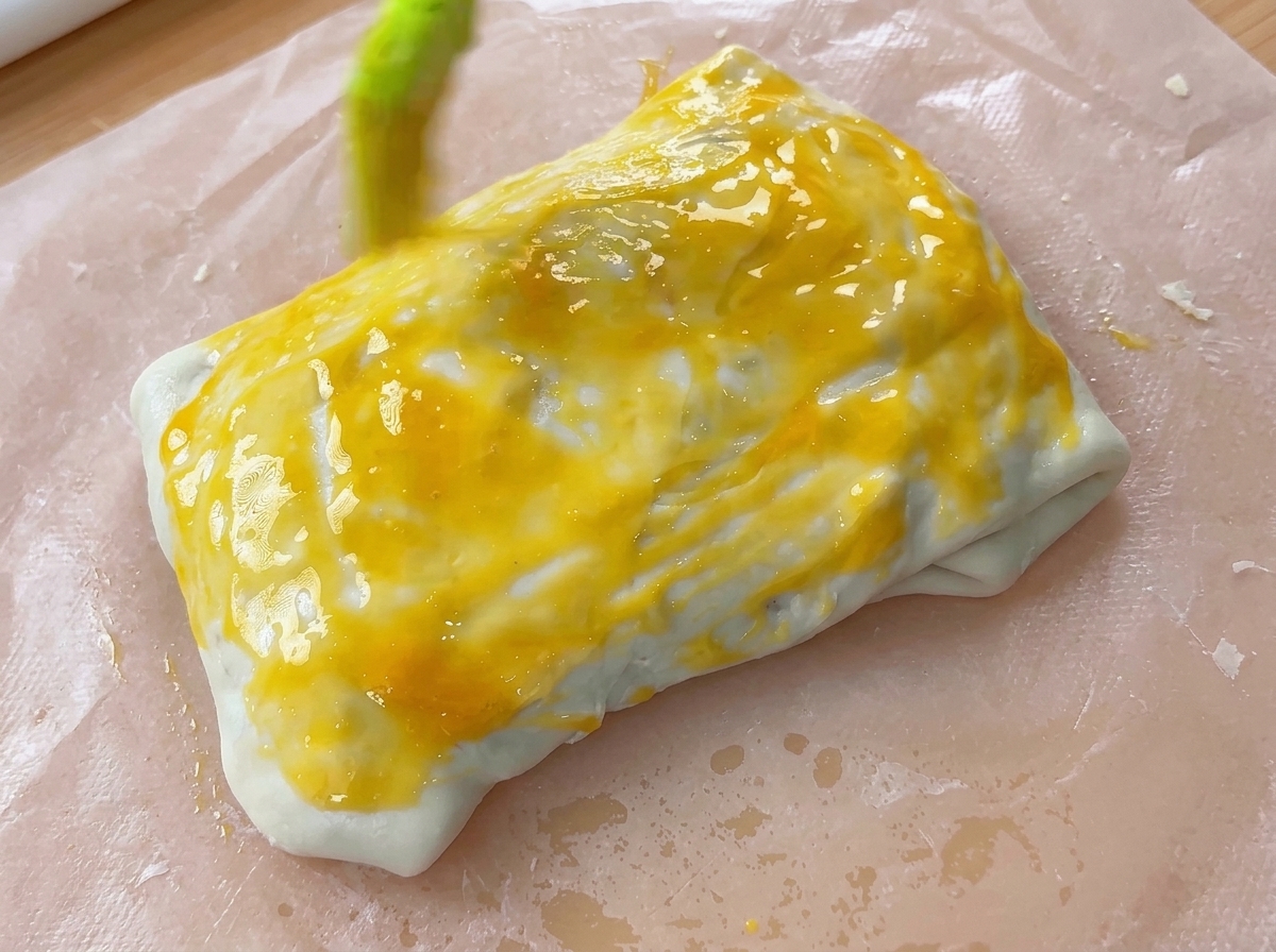 Un pennello da cucina che applica uno strato di tuorlo duovo giallo brillante su un fagottino di pasta chiuso.