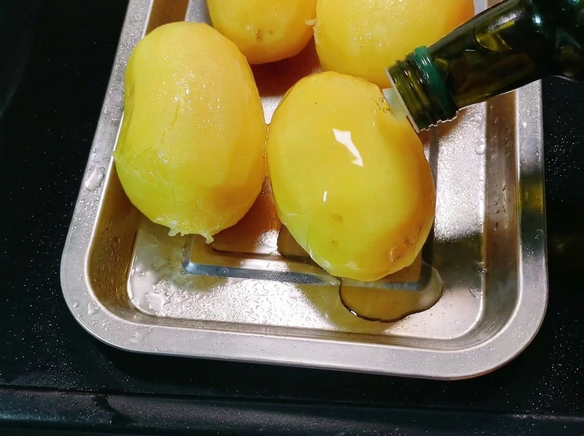 Olio da cucina versato da una bottiglia sulle patate pelate in una teglia da forno di metallo.