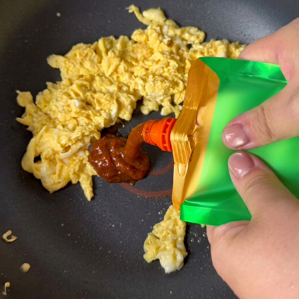 Pasta di soia gialla spremuta da una confezione verde sulle uova strapazzate in padella.