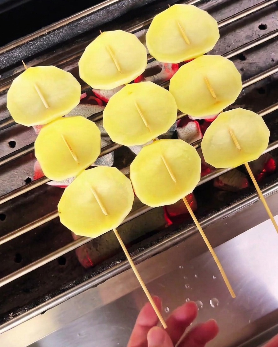 Fette di patate chiare infilzate sugli spiedini messe ad asciugare su un grill a carbonella.
