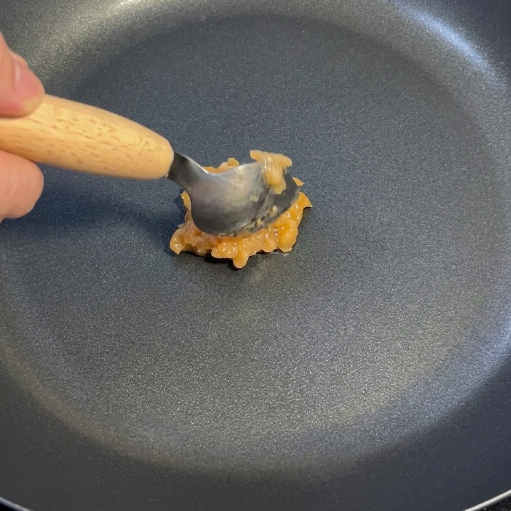 Un cucchiaio di salsa di aglio e zenzero mescolata in una padella antiaderente scura a fuoco basso.