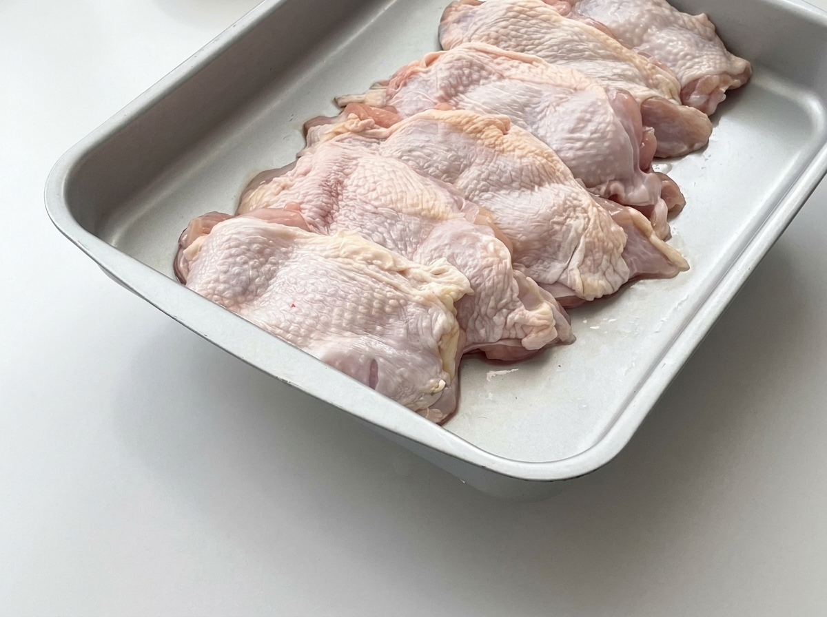 Cosce di pollo crude disposte in un vassoio di metallo con fette di zenzero fresco posizionate sopra.