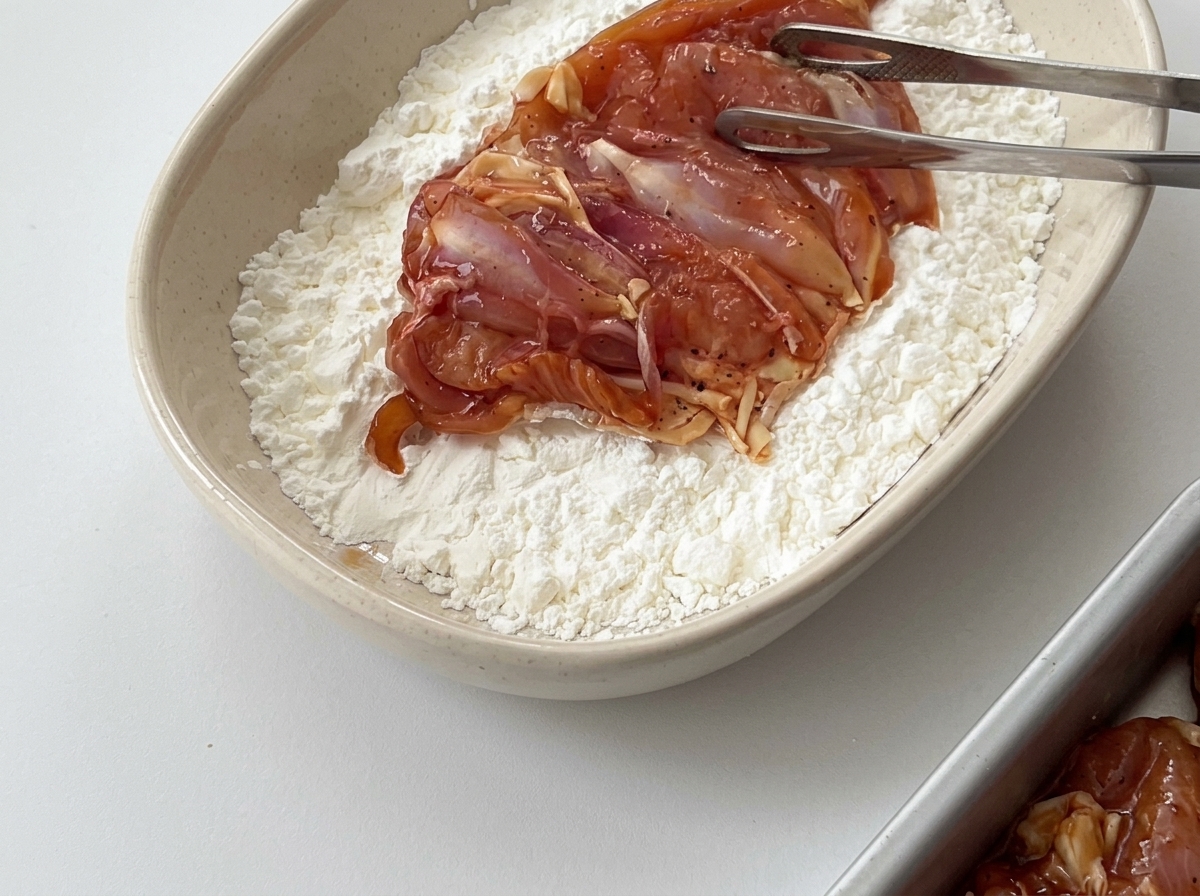 Un pezzo di pollo marinato preso con le pinze in una ciotola piena di amido di mais bianco.