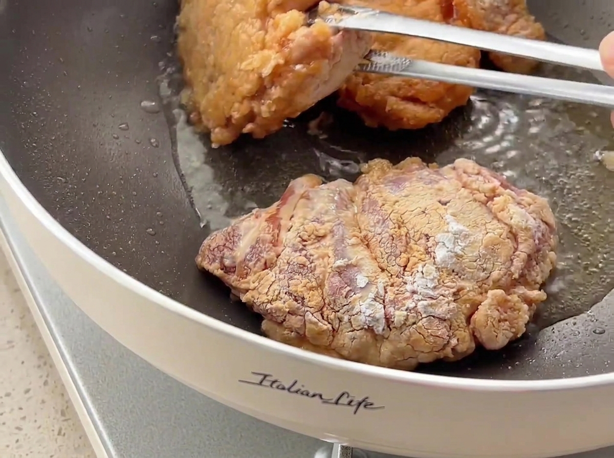 Pezzi di pollo girati con le pinze in una padella, che mostrano una crosta dorata e croccante.