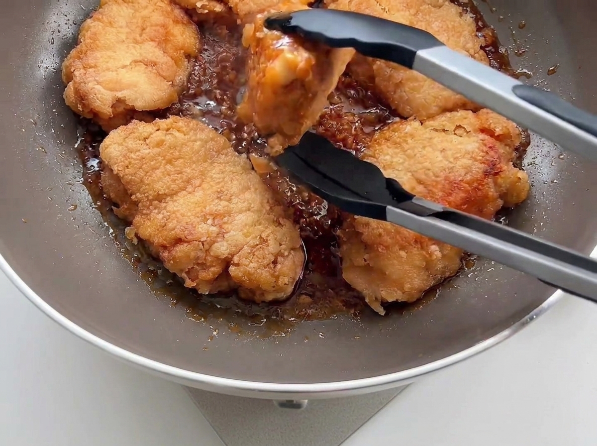 Pollo fritto croccante rimesso in padella con la salsa allaglio usando pinze da cucina nere.