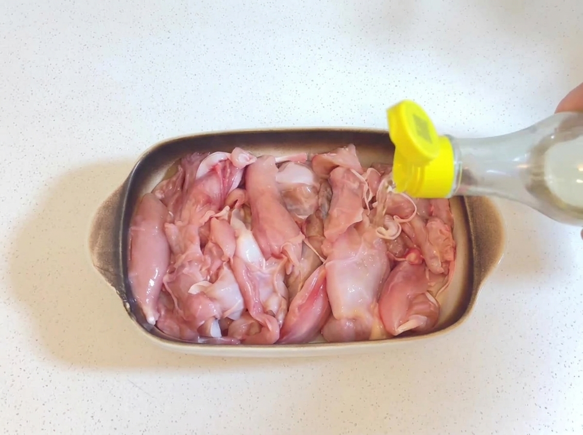 Vino da cucina trasparente versato da una piccola bottiglia con tappo giallo sui pezzi di pollo crudi in una teglia.