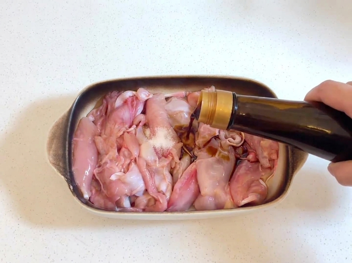Salsa di soia scura versata da una bottiglia di vetro sui pezzi di pollo crudi, con un piccolo pizzico di sale visibile sul pollo.