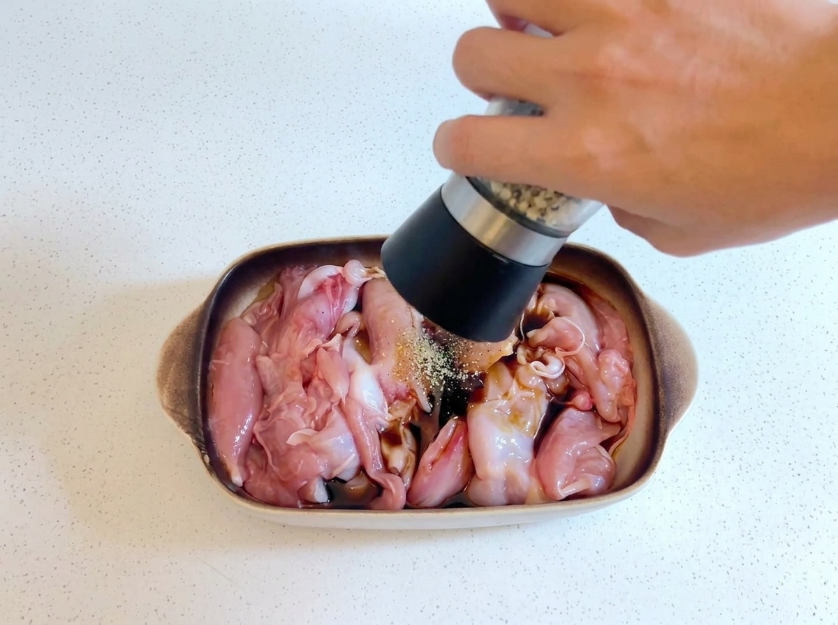 Una mano che tiene un macinapepe e macina pepe fresco sul pollo crudo in marinatura in un piatto.