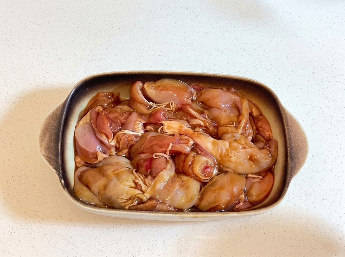 Una teglia rettangolare in ceramica riempita con pezzi di pollo marinati, strettamente coperta con pellicola trasparente.