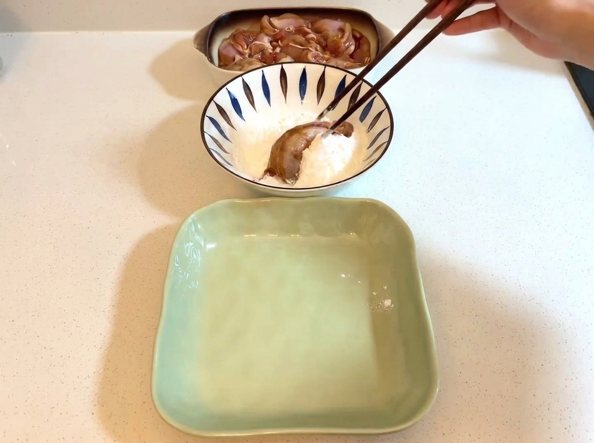 Bacchette che tengono un pezzo di pollo marinato allinterno di una ciotola riempita di amido bianco.