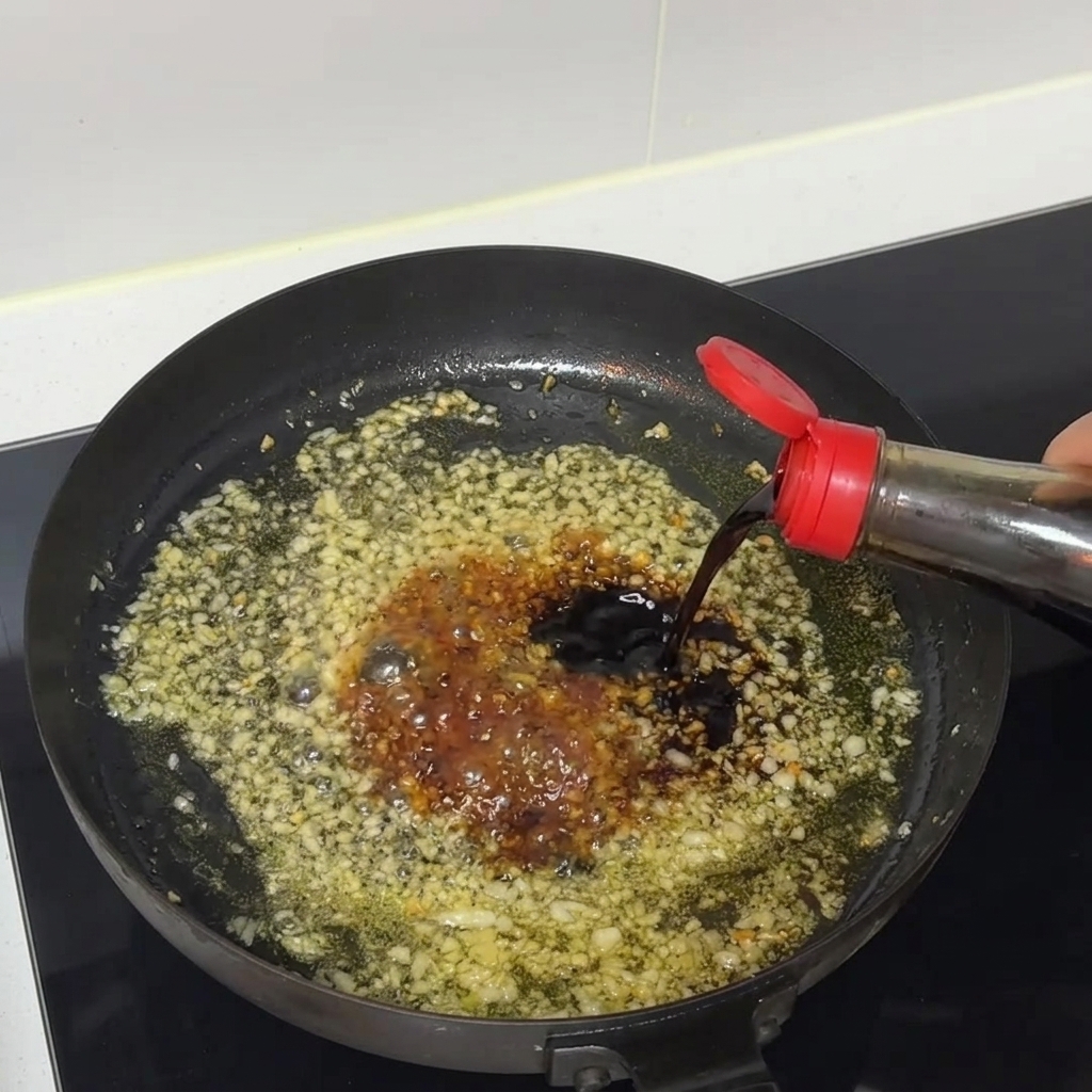Salsa di soia scura che cola da una bottiglia in una padella sfrigolante di aglio tritato e salsa.