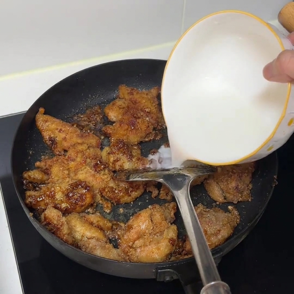 Acqua e amido versati da una ciotola bianca in una padella piena di pezzi di pollo cotti.