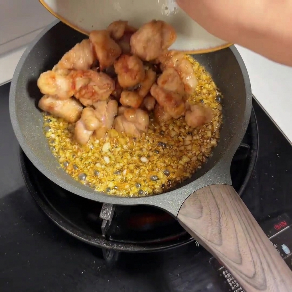 Pezzi di pollo pre-fritti che vengono versati da un piatto in una padella piena di salsa allaglio.