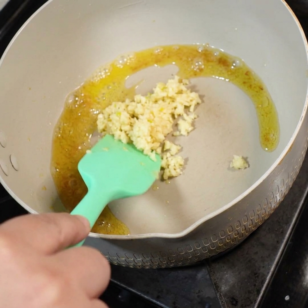 Aglio fresco tritato aggiunto al burro sciolto in una padella, mescolato con una spatola in silicone verde.