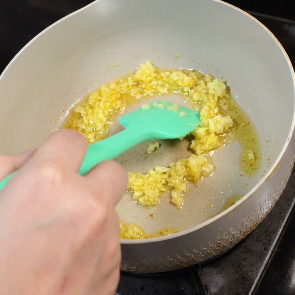 Aglio tritato che frigge nel burro sciolto in una padella, mescolato con una spatola in silicone verde.