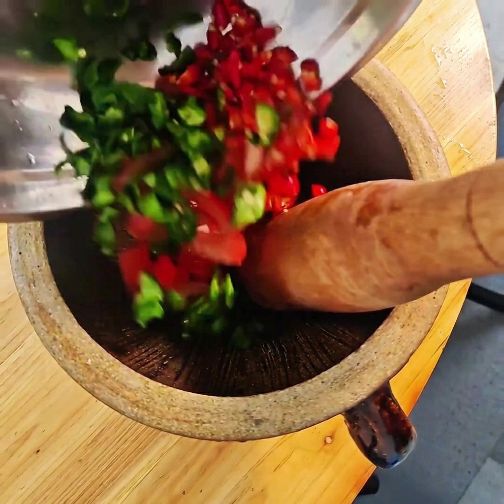 Peperoncini verdi e rossi tagliati e pomodori pestati in un mortaio di legno con un pestello.