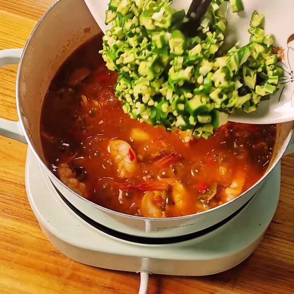Okra verde fresca tagliata fatta scivolare da un piatto bianco nello stufato bollente.