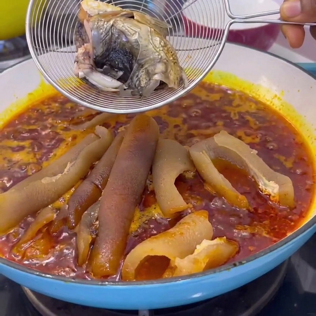Un colino a rete che tiene pezzi di pesce cotto abbassato in una pentola con salsa rossa che sobbolle e cotenna di maiale.