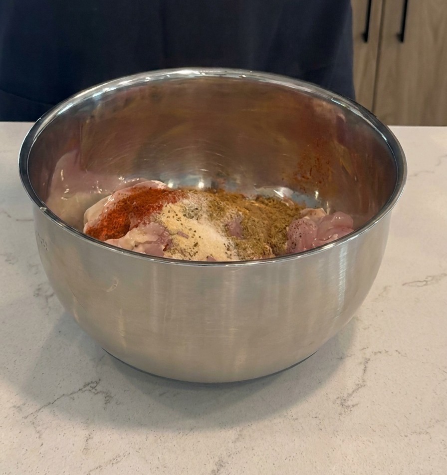 Aggiunta di marinatura rosso scuro e spezie secche alle cosce di pollo crude dentro una ciotola di miscelazione in acciaio inossidabile.