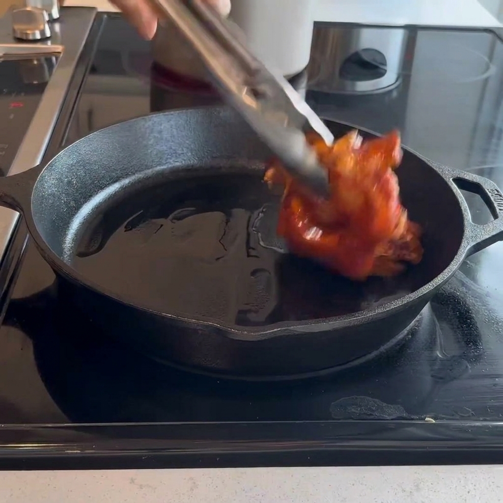 Cosce di pollo crude marinate messe con la pelle rivolta verso il basso in una padella di ghisa calda usando pinze da cucina.