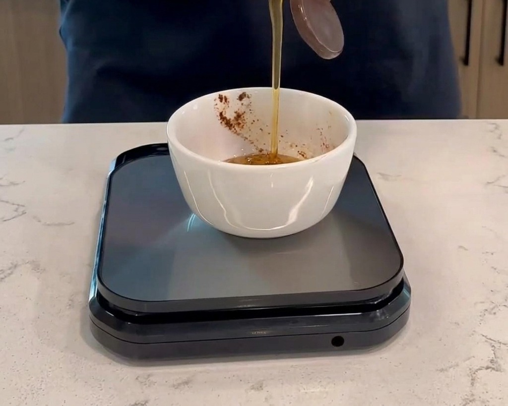 Versare il miele da una bottiglia di plastica in una ciotola bianca piccola con salsa rosso scuro, posizionata su una bilancia da cucina digitale nera.