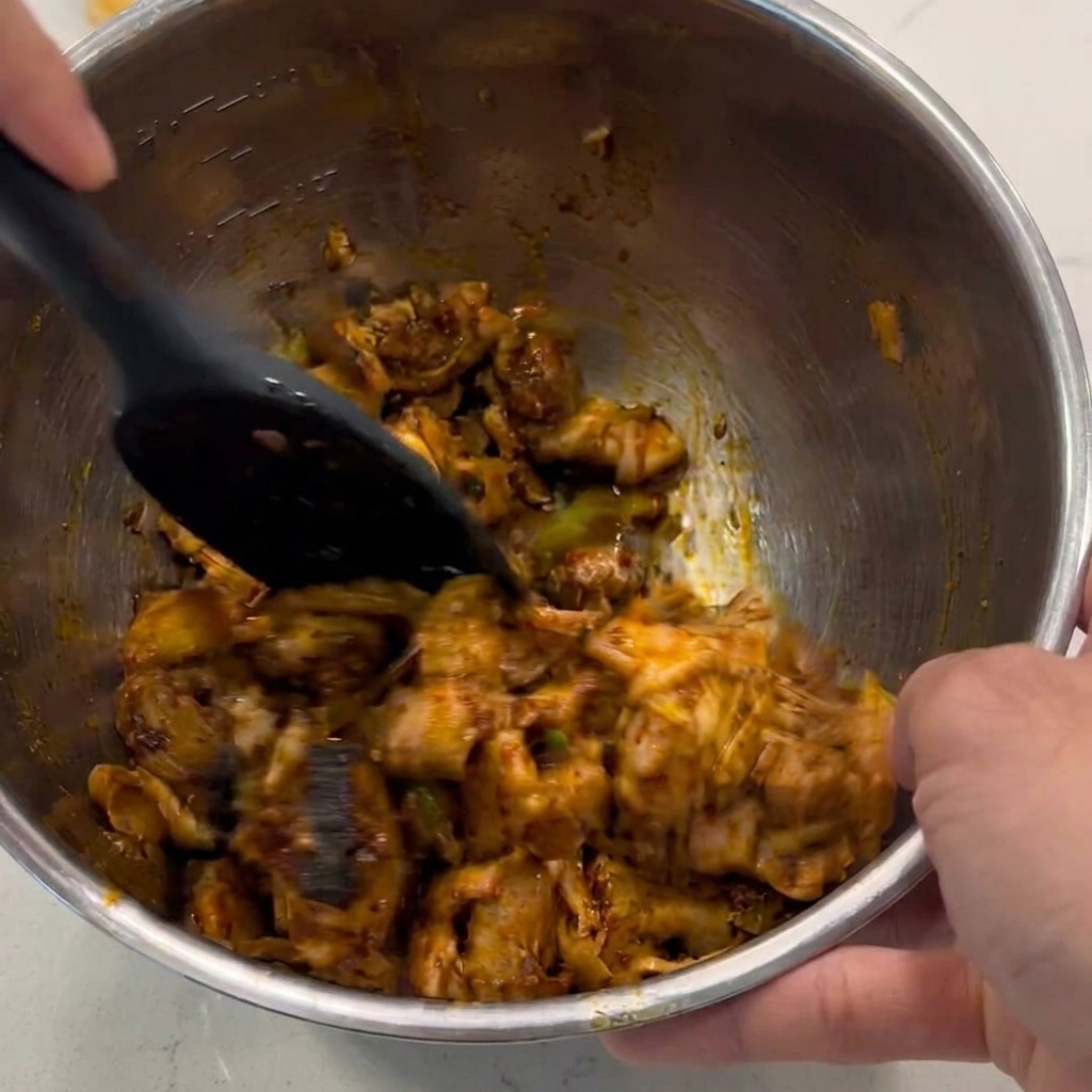 Una spatola nera che mescola e gira i pezzi di pollo a dadini per ricoprirli di una salsa marrone lucida dentro una ciotola di metallo.