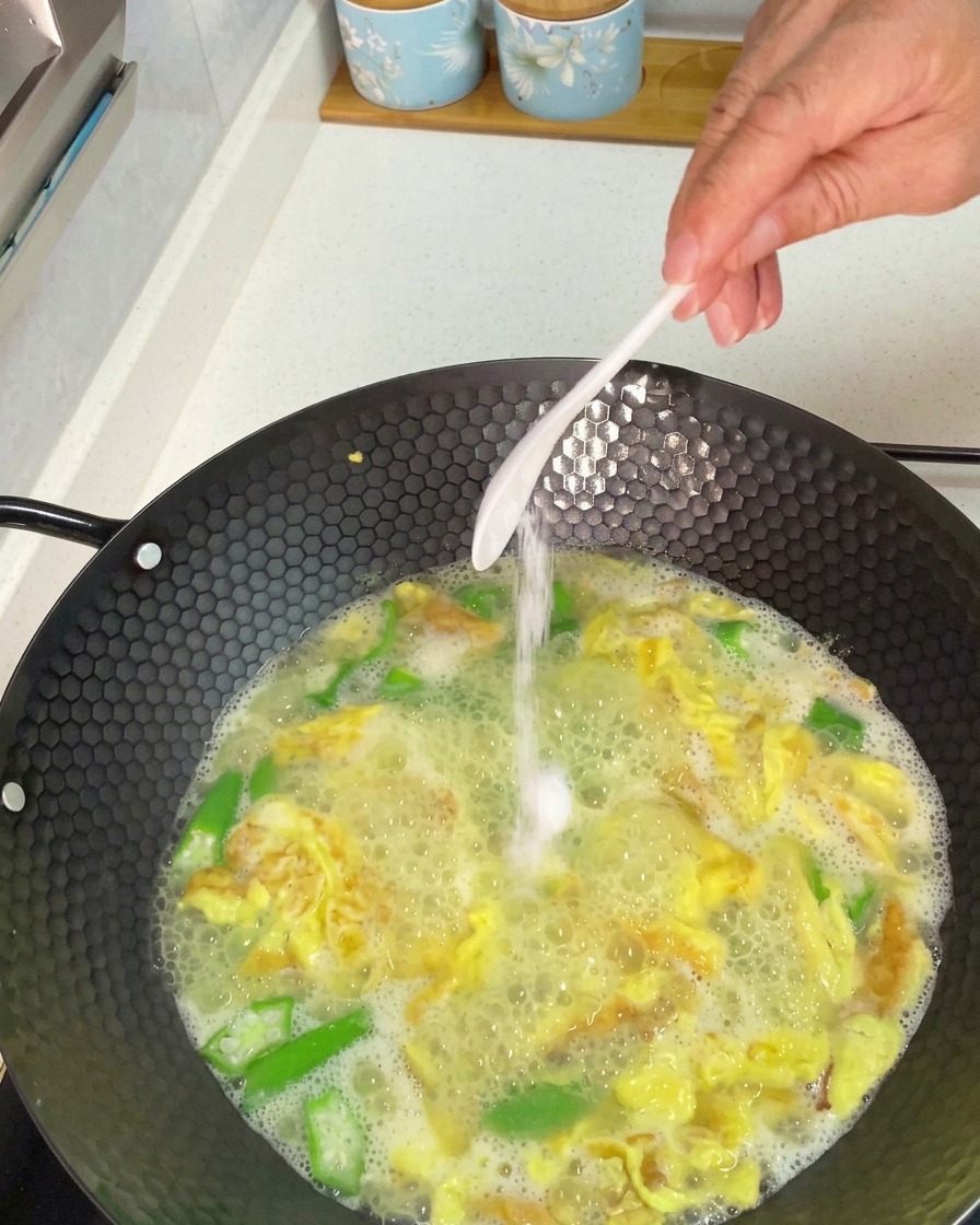 Mano che aggiunge un cucchiaio di sale bianco in un wok in ebollizione di zuppa di okra e uova.