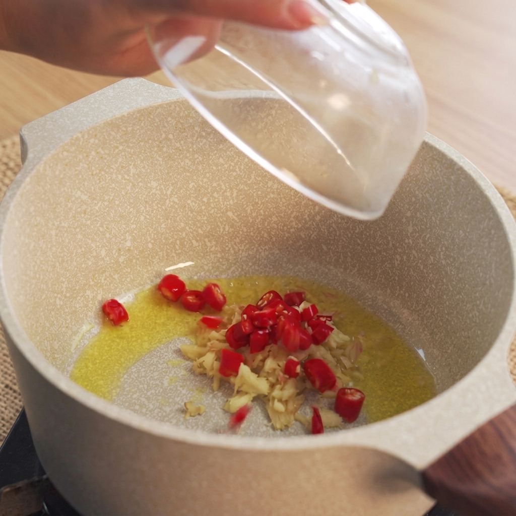 Mano che fa cadere peperoncini rossi tritati in una pentola con olio riscaldato e zenzero tritato.