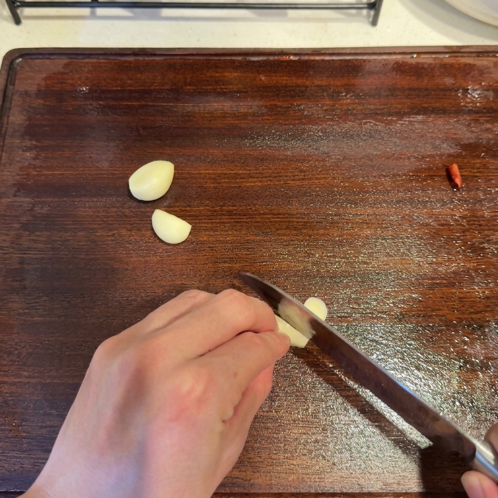 Spicchi daglio affettati con un coltello su un tagliere di legno accanto a un pezzo di peperoncino rosso.